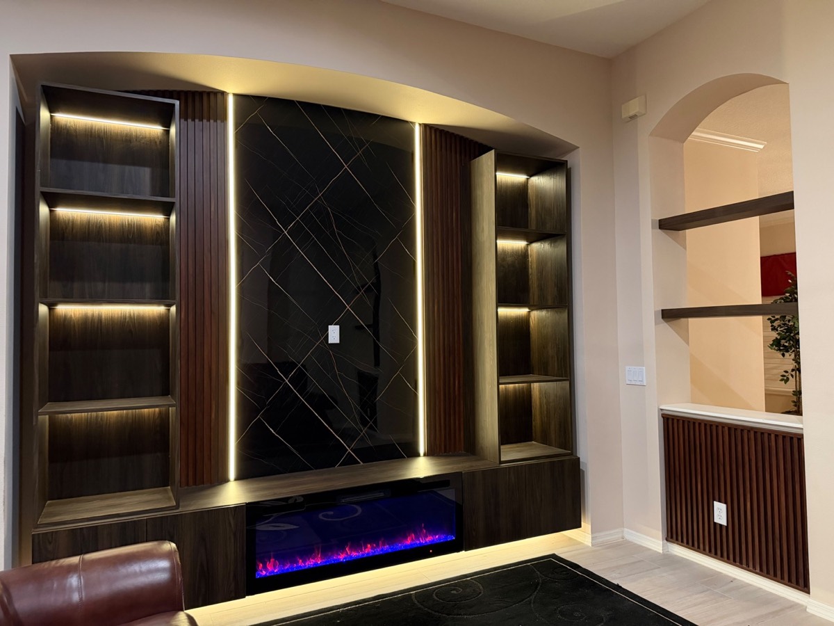 Dark built-in cabinetry with lit shelving around a TV and electric fireplace